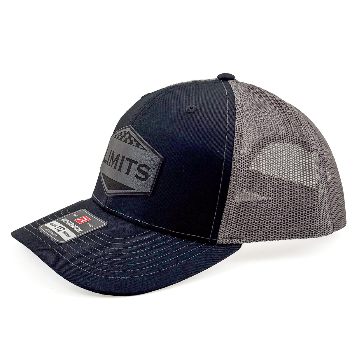 A Richardson 112 trucker hat in black and charcoal grey mesh, featuring a black hexagonal patch on the front with the word 'LIMITS' and a Richardson brand sticker on the visor.