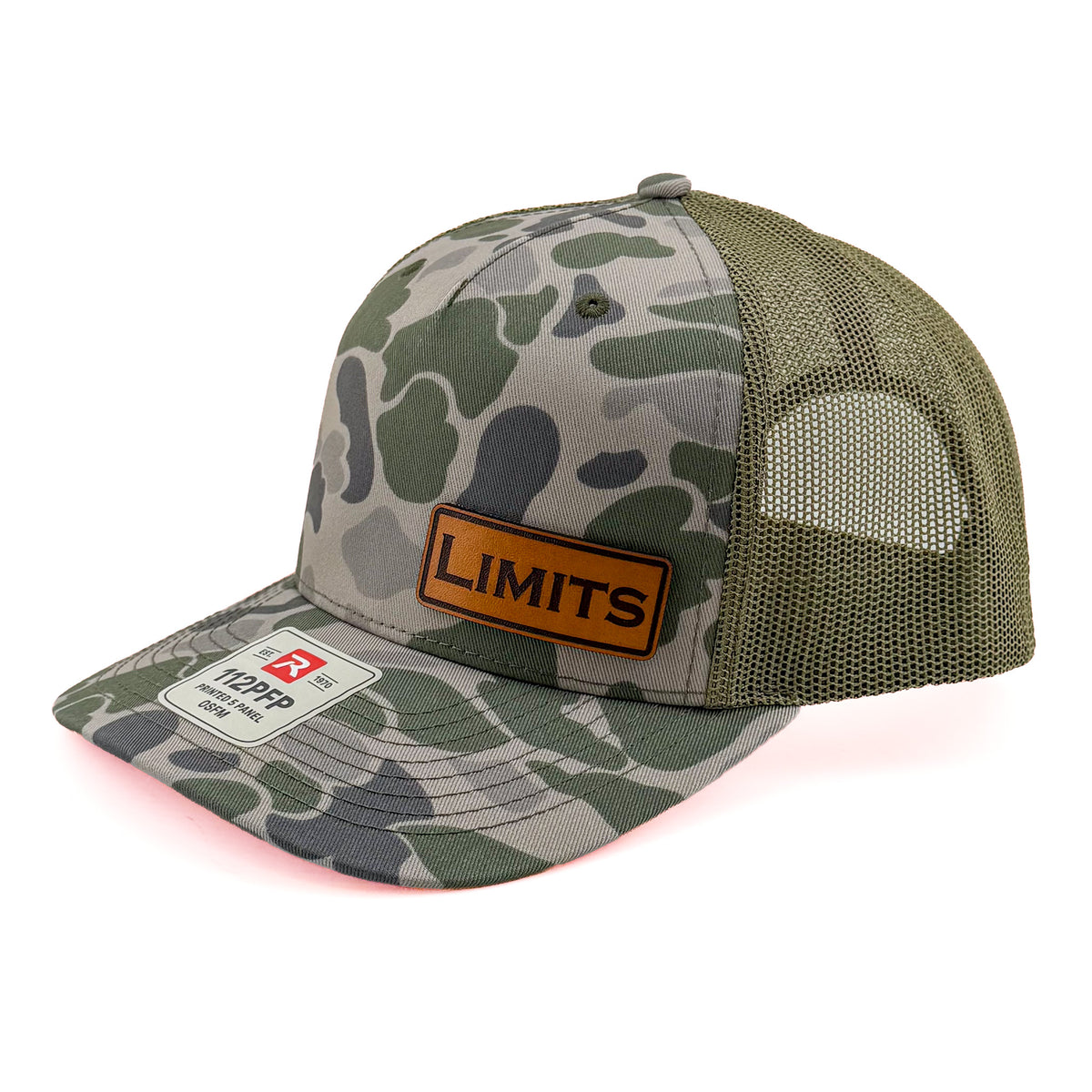 A camouflage trucker hat with green and gray camo print on the front panel and brim, olive mesh back panels, and a brown leather patch on the front with the brand name 'LIMITS'.