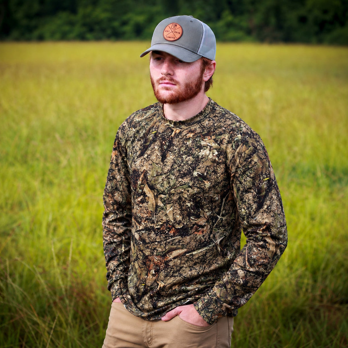 A man wearing a long-sleeve camouflage hunting shirt and a gray cap stands in a grassy field.