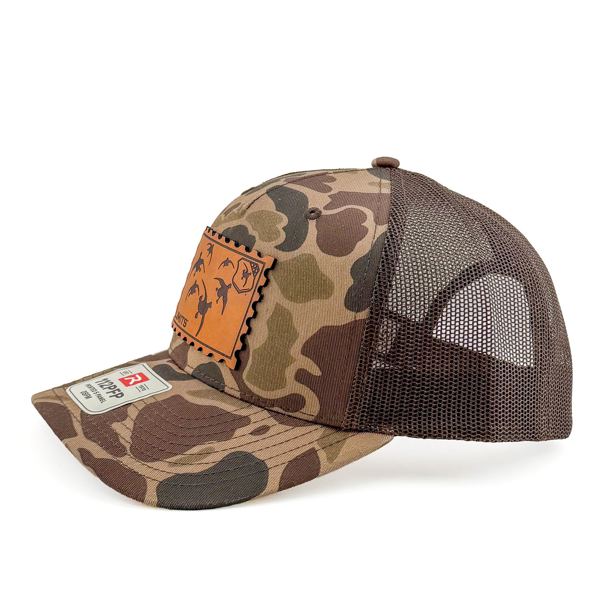 A camouflage trucker hat with a brown mesh back, featuring a tan leather patch on the front with a duck silhouette and a size tag on the brim.