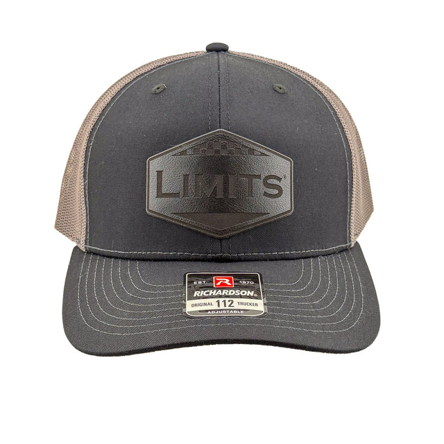 A Richardson 112 trucker hat in black and charcoal grey mesh, featuring a black hexagonal patch on the front with the word 'LIMITS' and a Richardson brand sticker on the visor.