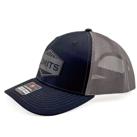 A Richardson 112 trucker hat in black and charcoal grey mesh, featuring a black hexagonal patch on the front with the word 'LIMITS' and a Richardson brand sticker on the visor.