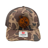 A camouflage trucker hat with brown mesh back panels and a circular brown leather patch on the front. The Limits patch features a duck silhouette and the text 'WE DEAL IN STEEL'.
