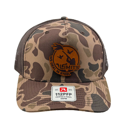 A camouflage trucker hat with brown mesh back panels and a circular brown leather patch on the front. The Limits patch features a duck silhouette and the text 'WE DEAL IN STEEL'.