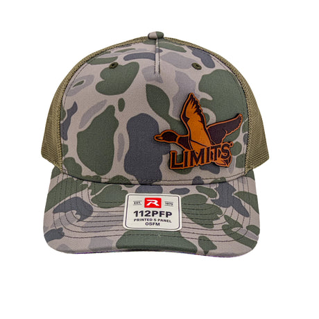 A camouflage trucker hat with olive green mesh back panels and a front patch featuring a leather duck logo and the word 'LIMITS'. A white size and material label is visible on the brim.