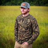 A man wearing a long-sleeve camouflage hunting shirt and a gray cap stands in a grassy field.