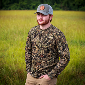 A man wearing a long-sleeve camouflage hunting shirt and a gray cap stands in a grassy field.
